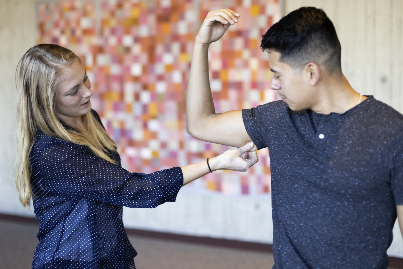 A dietitian (female, blonde hair and light skin) is checking the skin fold of an arm on a male patient. He has dark hair and light skin. His elbow is making an L shape as he watches her pinch the underfold of his arm