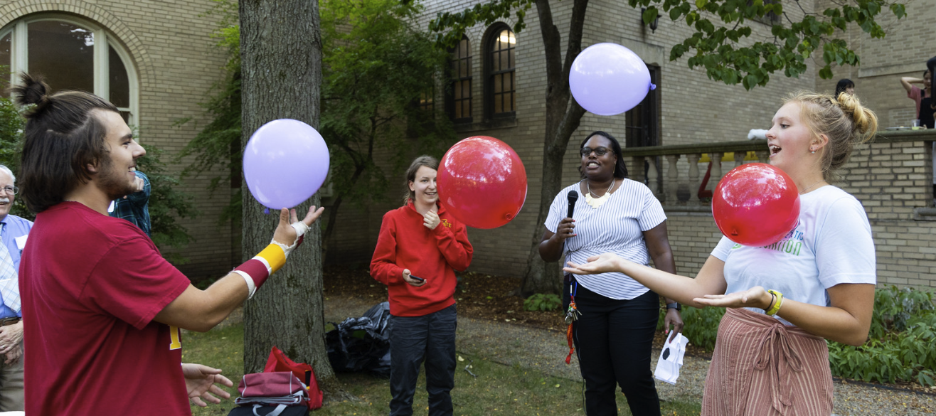 Four people outside playing with red and purple balloons. There are three college-age women and one male.