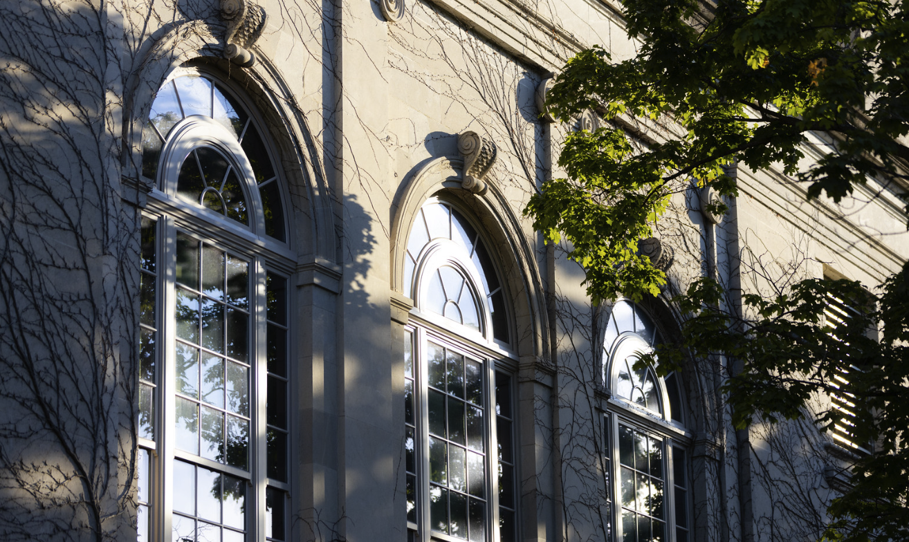 Three arched windows that are each two stories tall on a light, limestone building with green ivy growing on it