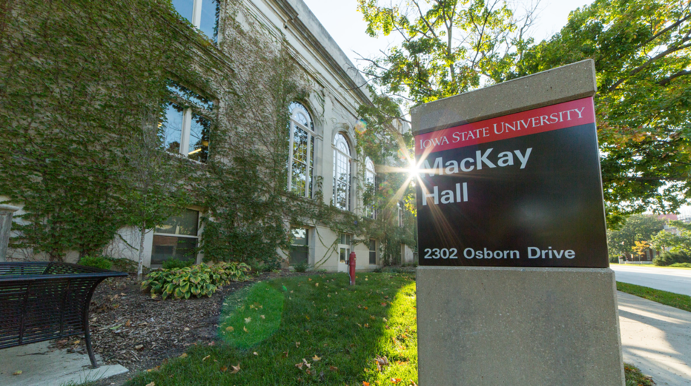 A building that has green vines covering it with a sign that says "MacKay Hall" - the sun is peeking between the sign and the building.